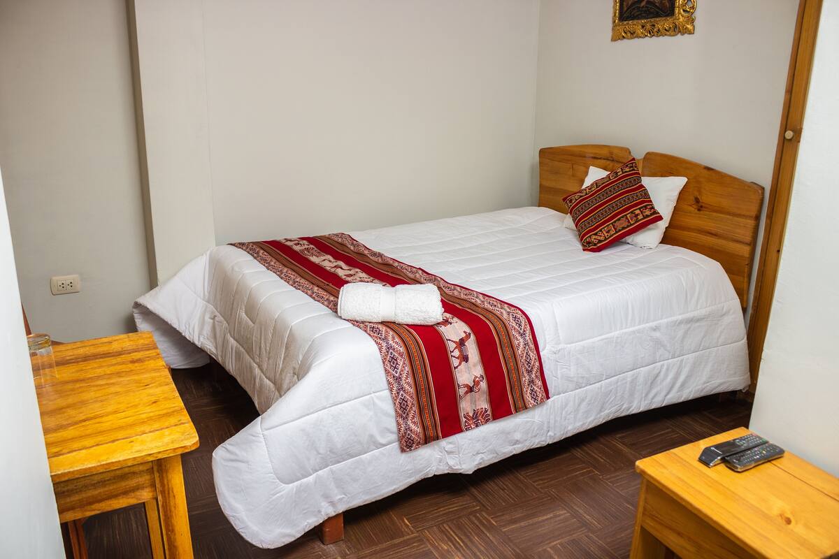A neatly made bed with a white comforter and a red and brown patterned blanket, a wooden headboard, a wooden nightstand with a remote control, and a small table with a glass container.