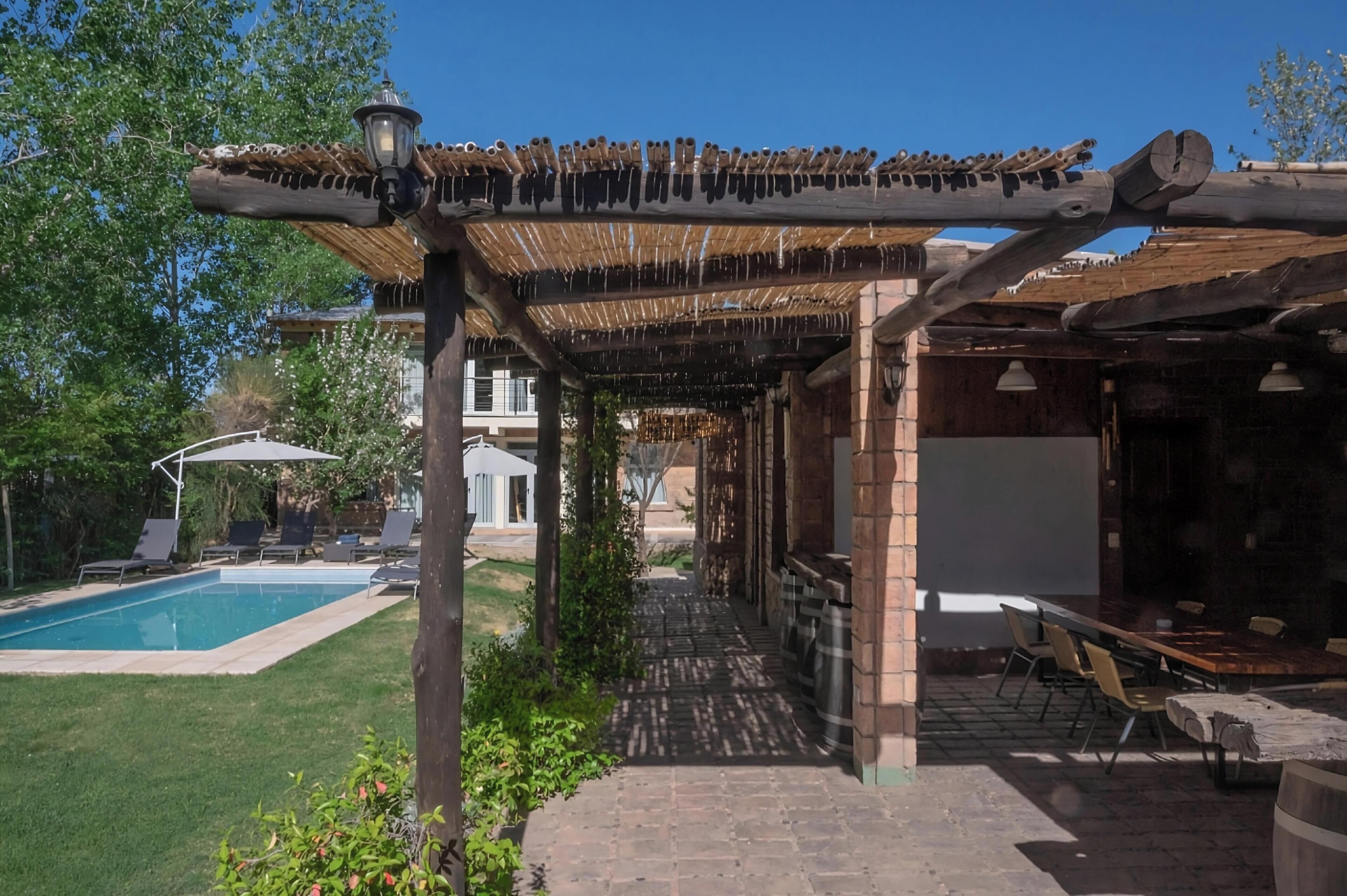 A covered outdoor area with a pool, lounge chairs, and a pergola with hanging lights.