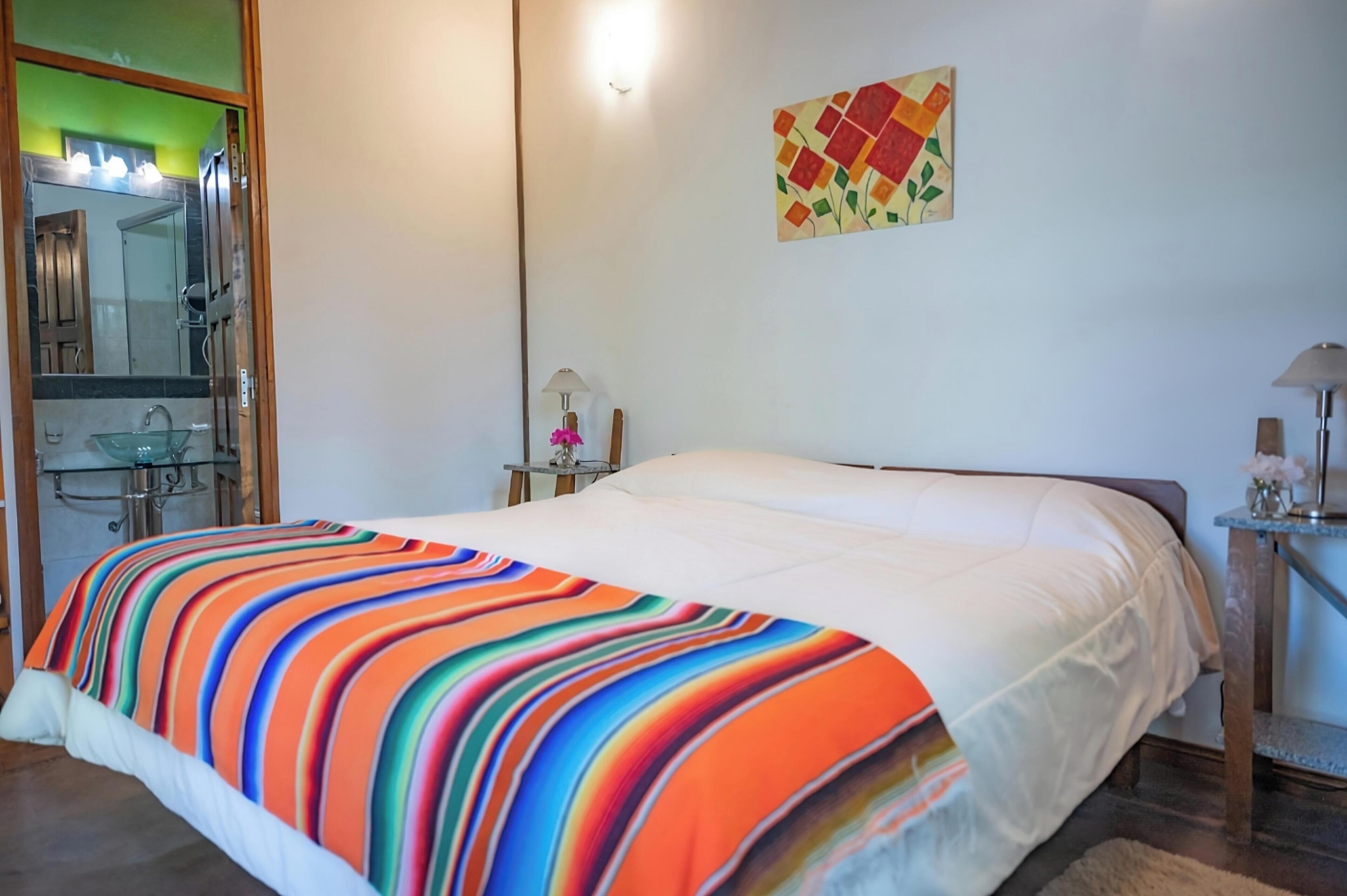 Un dormitorio con una cama grande, una mesita de noche con lámpara, un espejo y un baño visible a través de un mamparo de vidrio.