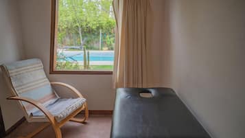 Relax in comfort with a view of the serene pool and lush greenery from this room's window, featuring a cozy seating area and a massage table.