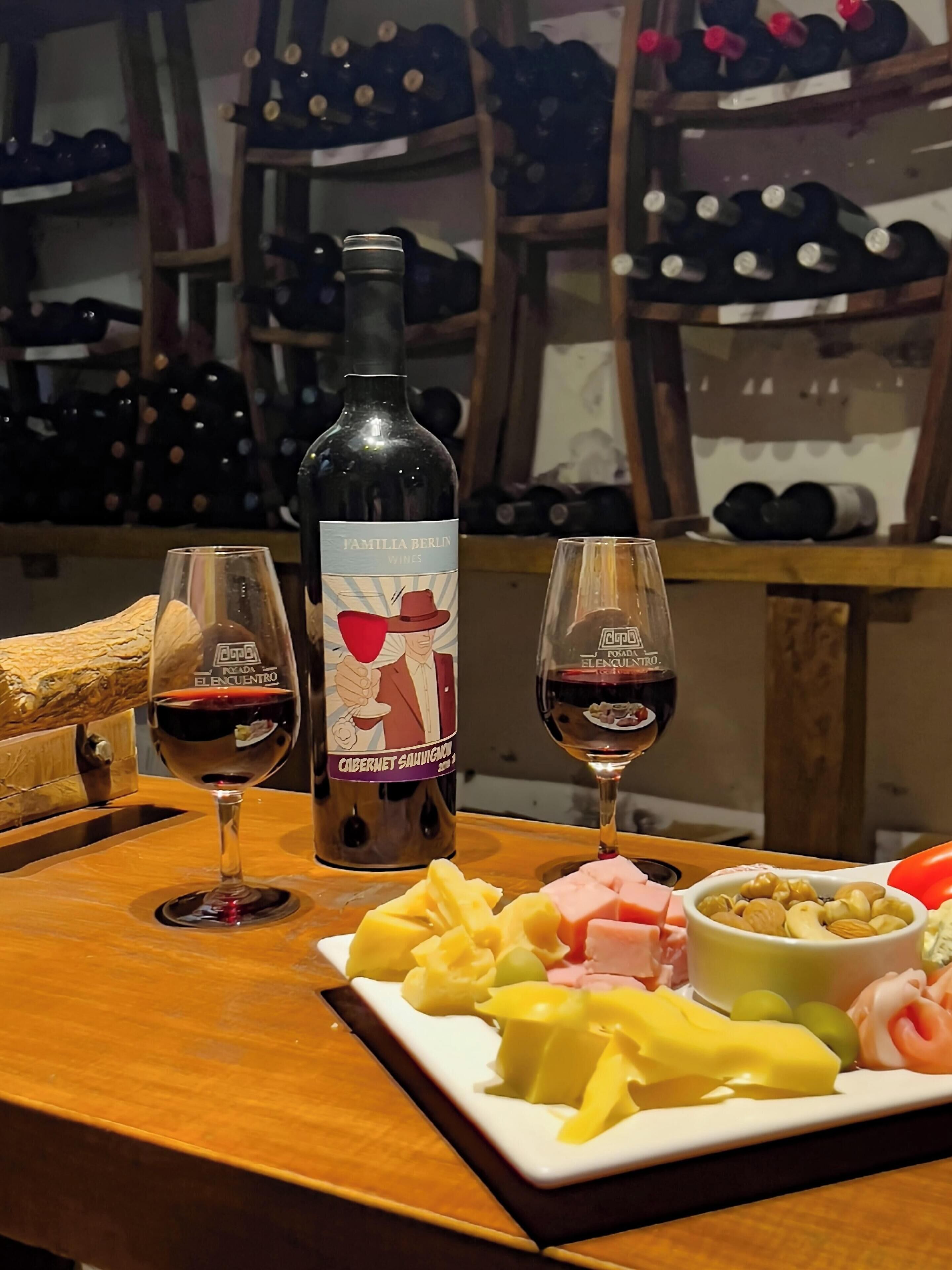 Et vinforsmagning-setup med en flaske Familia Berlin vin, to glas rødvin og en tallerken med ost og nødder.
