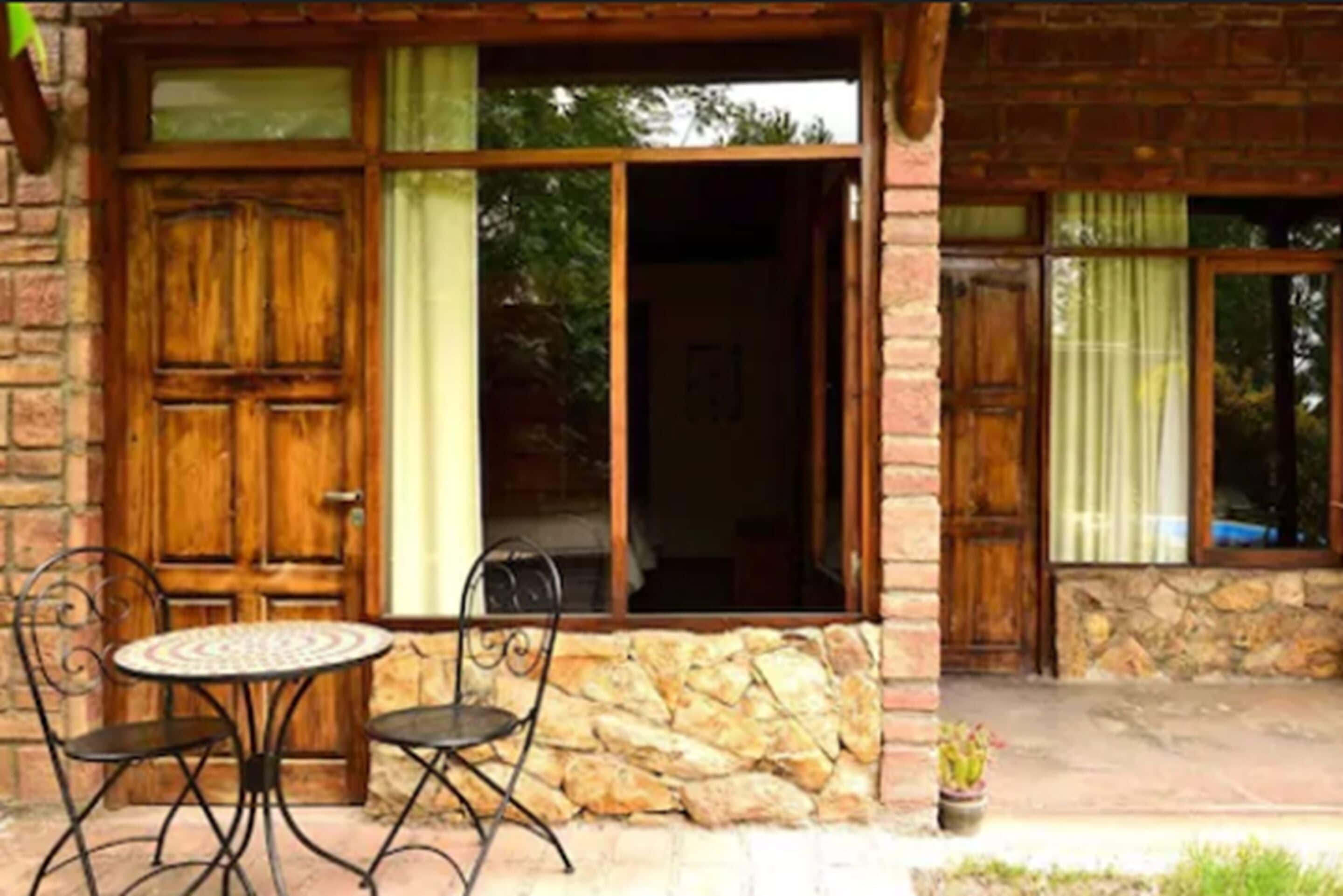 Fachada de piedra con puerta y ventanas de madera, una pequeña mesa redonda con sillas, y una planta en maceta en el exterior.