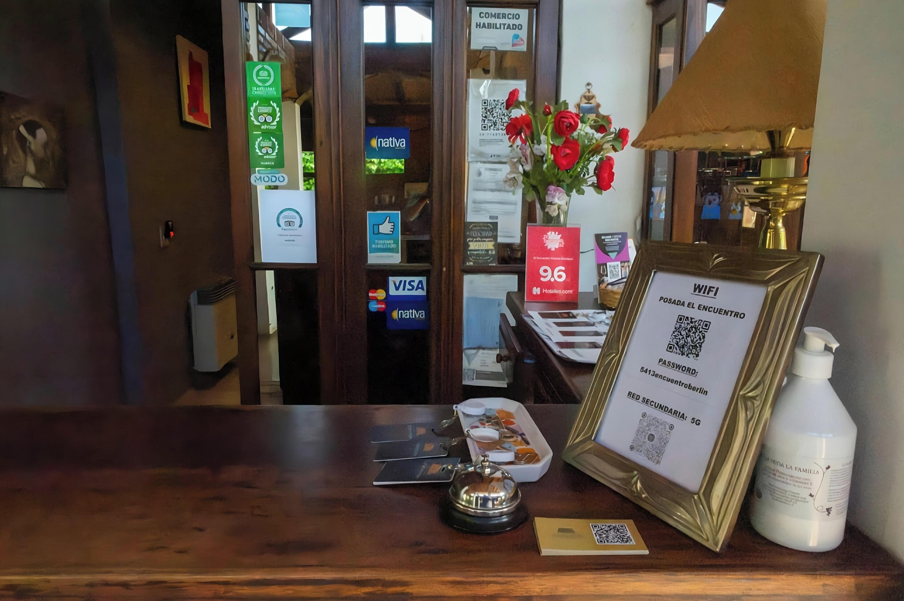 A hotel reception desk with a framed WiFi sign, a vase with red flowers, a lamp, and a bottle of hand sanitizer.