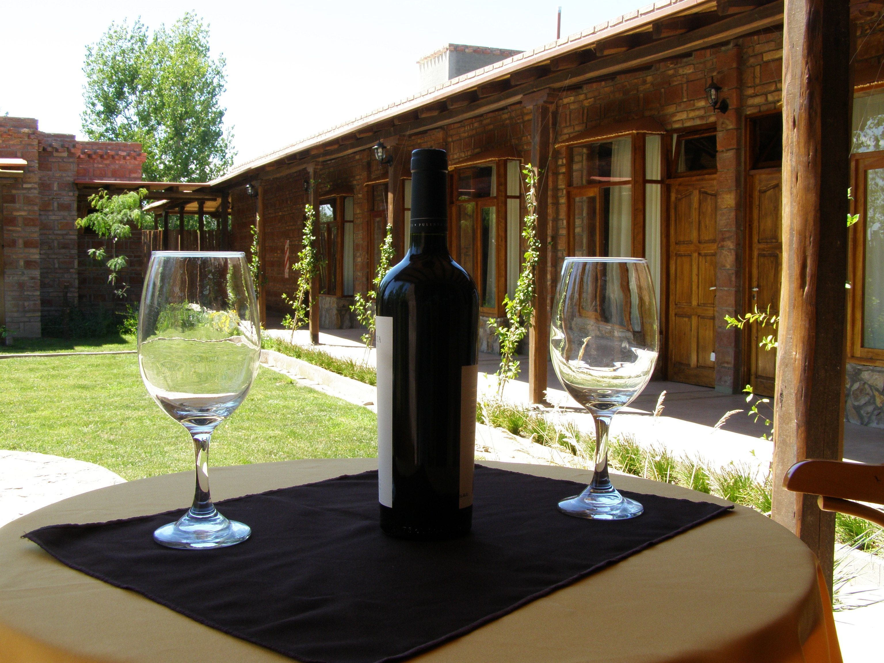 A bottle of wine on a table with two glasses, set outdoors in front of a brick building with wooden doors and windows.