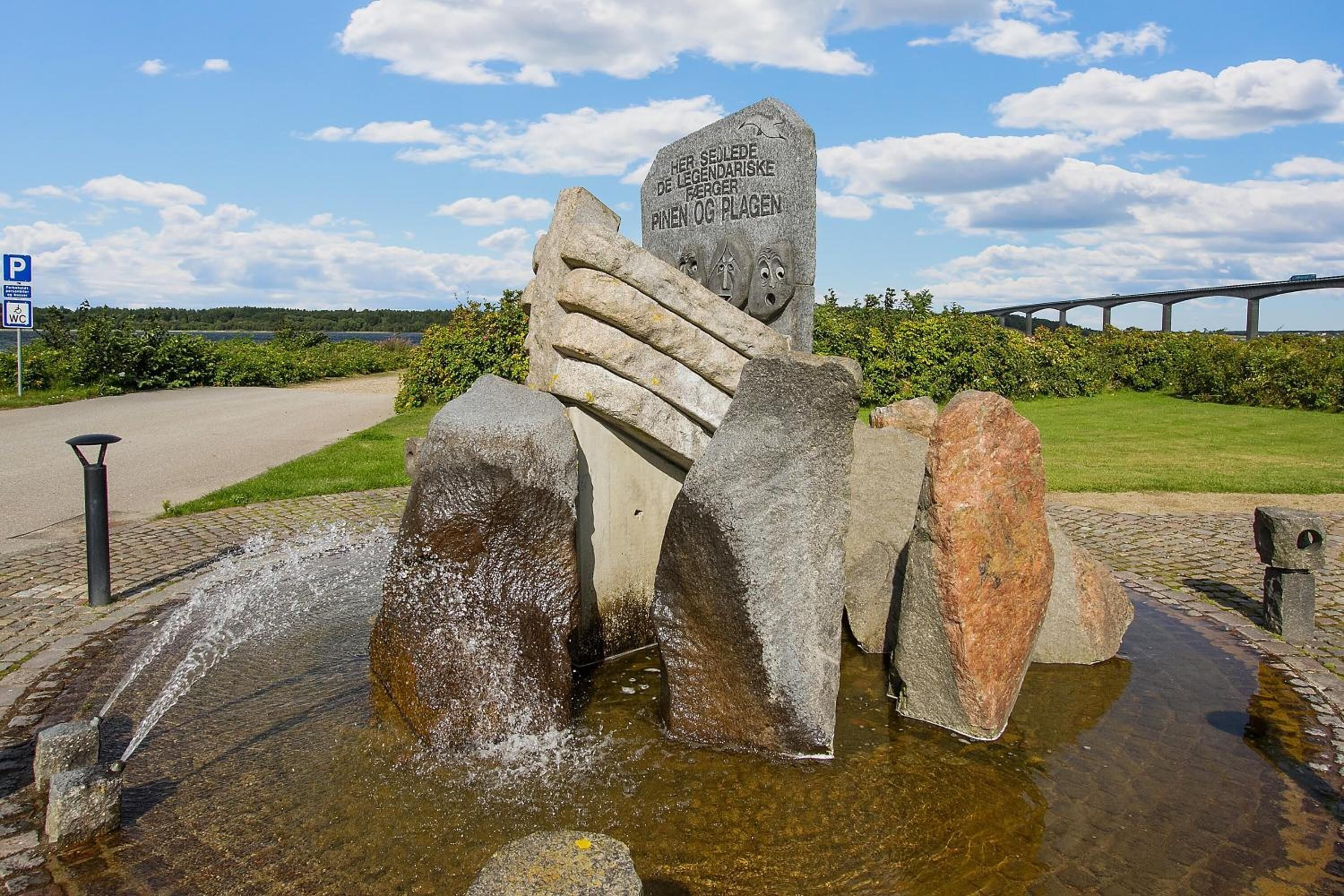Eine Steinskulptur mit einer Gedenktafel, gelegen in einer Parkanlage mit einem gepflasterten Weg und einer Brücke im Hintergrund.