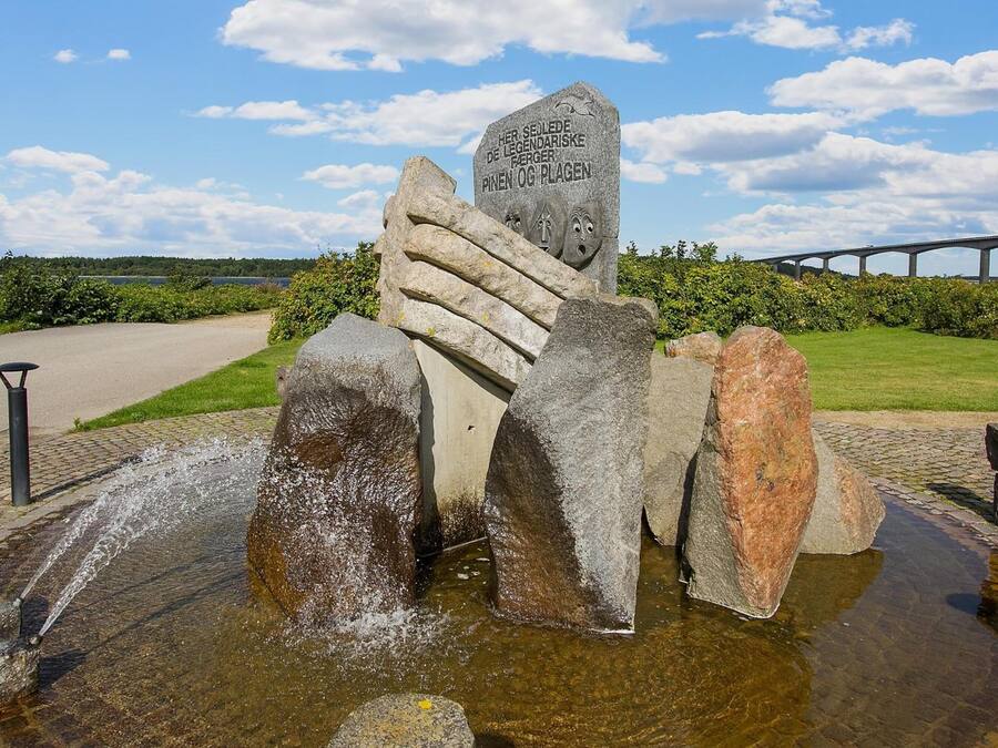 Una escultura de piedra con una placa en un parque, un sendero pavimentado y un puente al fondo.