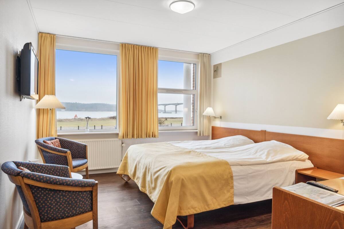 A hotel room with a bed, two chairs, a TV, and a window with curtains.