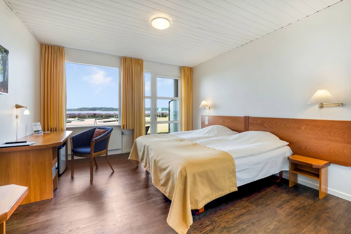 A hotel room with a bed, desk, chair, and a view of the outdoors.