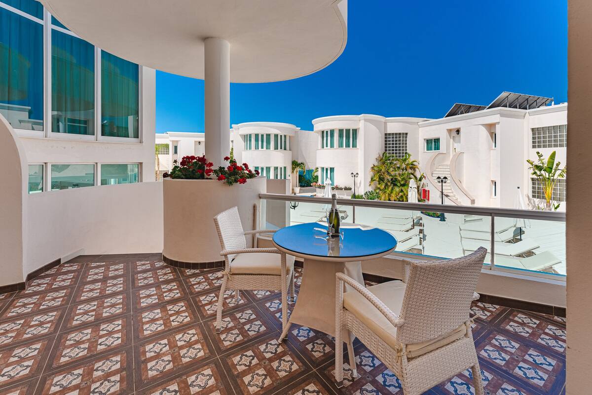 Ein Balkon mit einem runden Tisch und Korbstühlen, mit Blick auf ein Schwimmbad und weiße Gebäude.