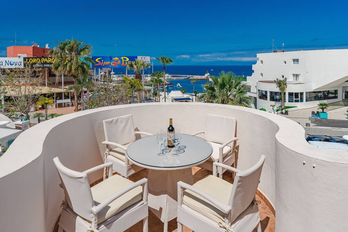 Ein runder Tisch mit vier weißen Stühlen, einer Champagnerflasche und mehreren Gläsern auf einer Terrasse mit Blick auf einen Strand und einen Hafen.