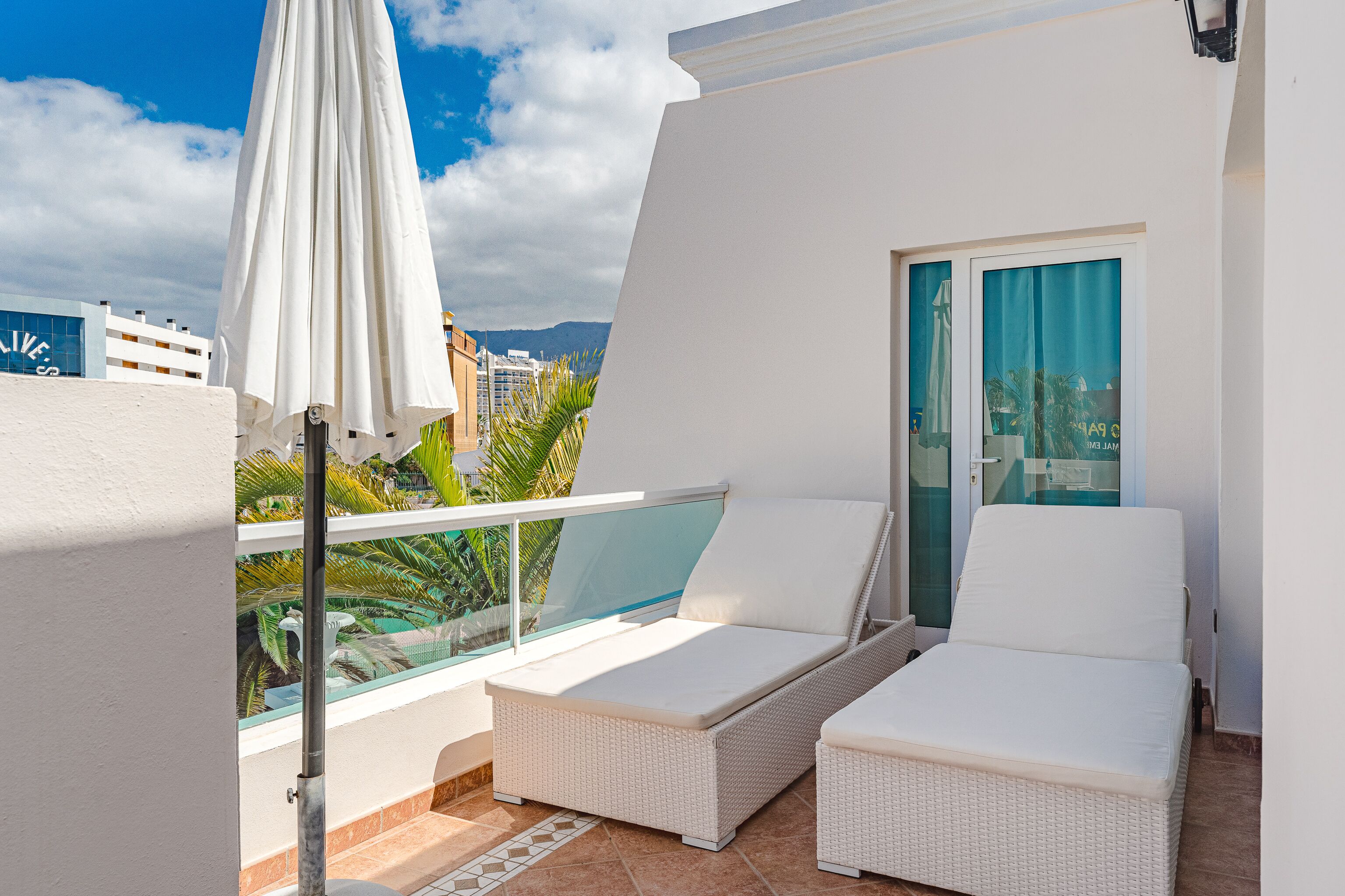 Un balcon avec deux fauteuils de relaxation en osier blanc, un parasol blanc et une porte vitrée donnant sur une piscine.