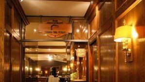 Um corredor estreito com painéis de madeira e uma placa indicando um restaurante. Há cadeiras ao longo das paredes e uma mesa ao longe.