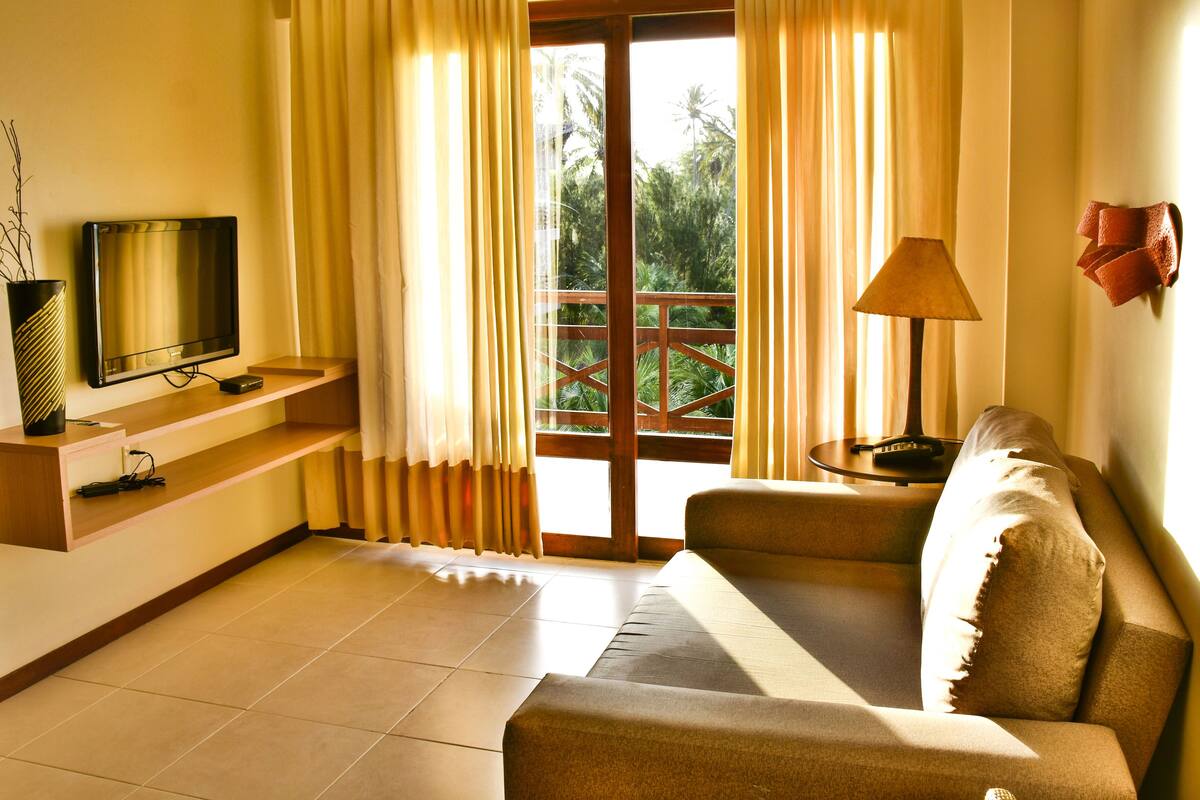 Una sala de estar con un sofá, un televisor sobre un mueble bajo, una lámpara y vistas al exterior con vegetación.