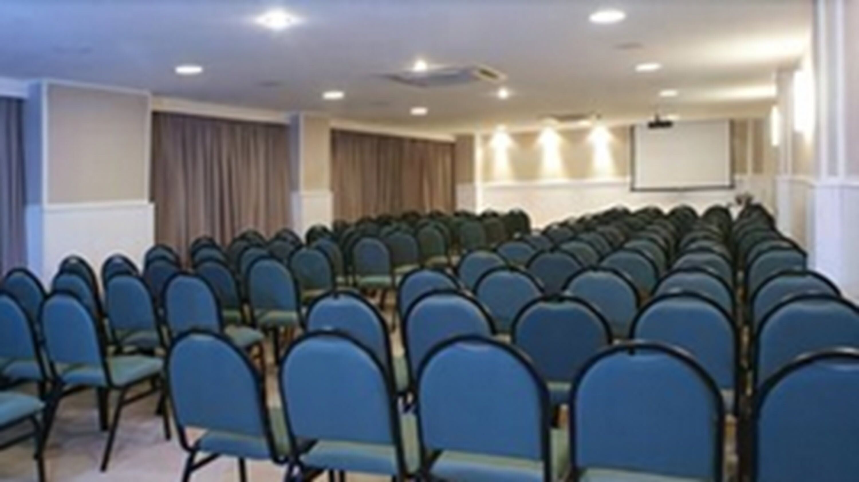 Un salón de conferencias con filas de sillas azules, una pizarra y una pantalla de proyector.