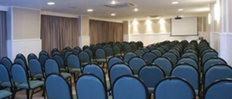 Un salón de conferencias con filas de sillas azules, una pizarra y una pantalla de proyector.