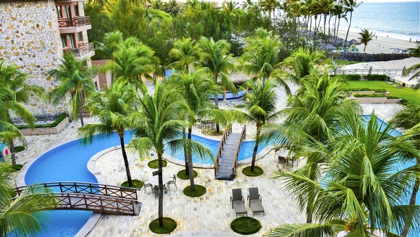 Zona de piscina en un resort, con palmeras, un puente y vistas a la playa.