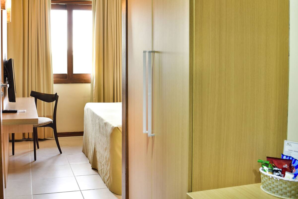 Habitación de hotel con cama, escritorio con silla, ventana con cortinas y armario de madera.
