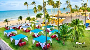 Un área de comedor al aire libre con mesas y sillas, rodeada de palmeras y cabañas de playa.
