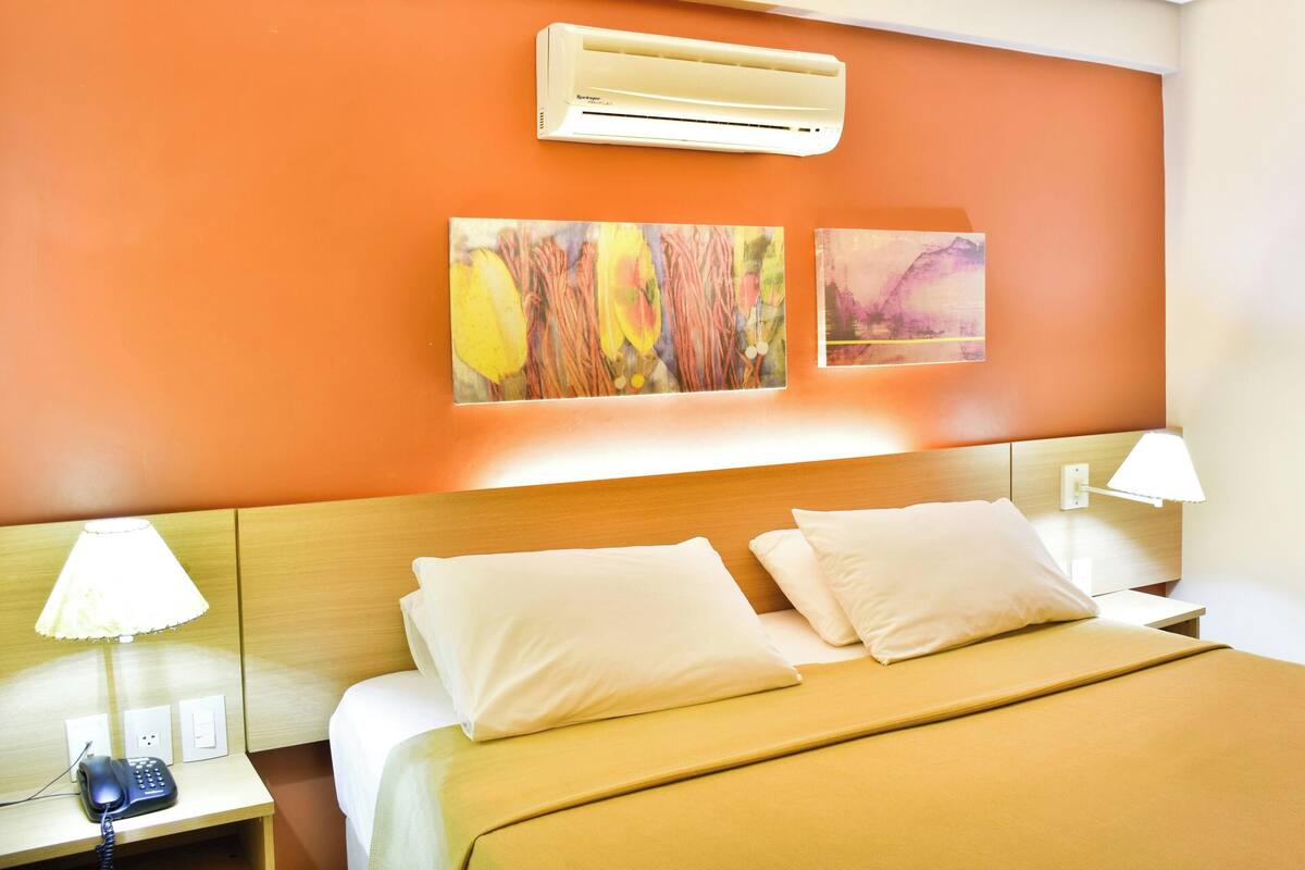 Una habitación de hotel con cama, lámparas de noche, aire acondicionado y una obra de arte en la pared.