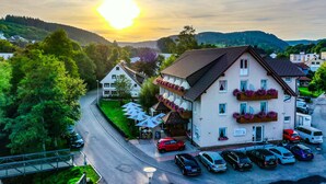 Un hotel con fachada blanca, varios balcones con jardineras y un estacionamiento con varios coches, y vistas al entorno verde y las colinas.