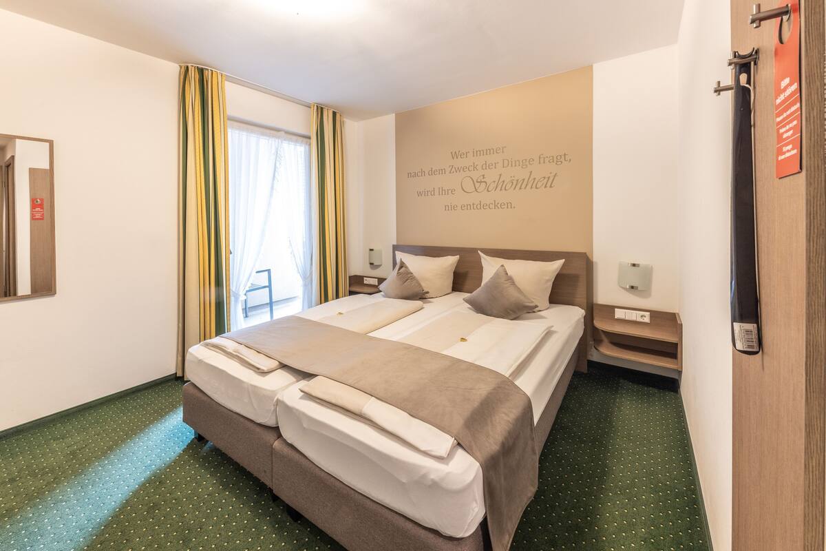 A hotel room with a large bed, two pillows, a bedside table, a nightstand, a wall-mounted light, a window with curtains, and a motivational quote on the wall.