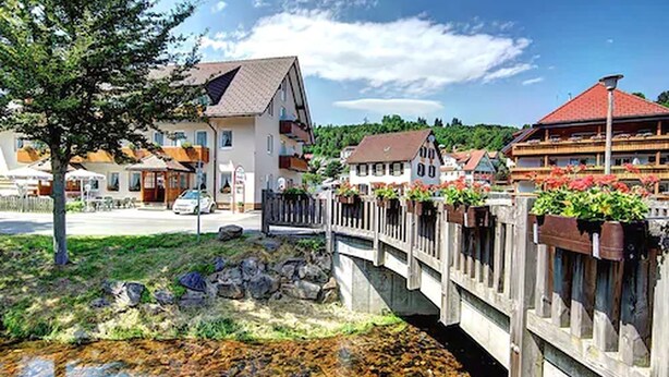 Vista a la orilla del río con casas tradicionales, un puente de madera y cielo despejado.