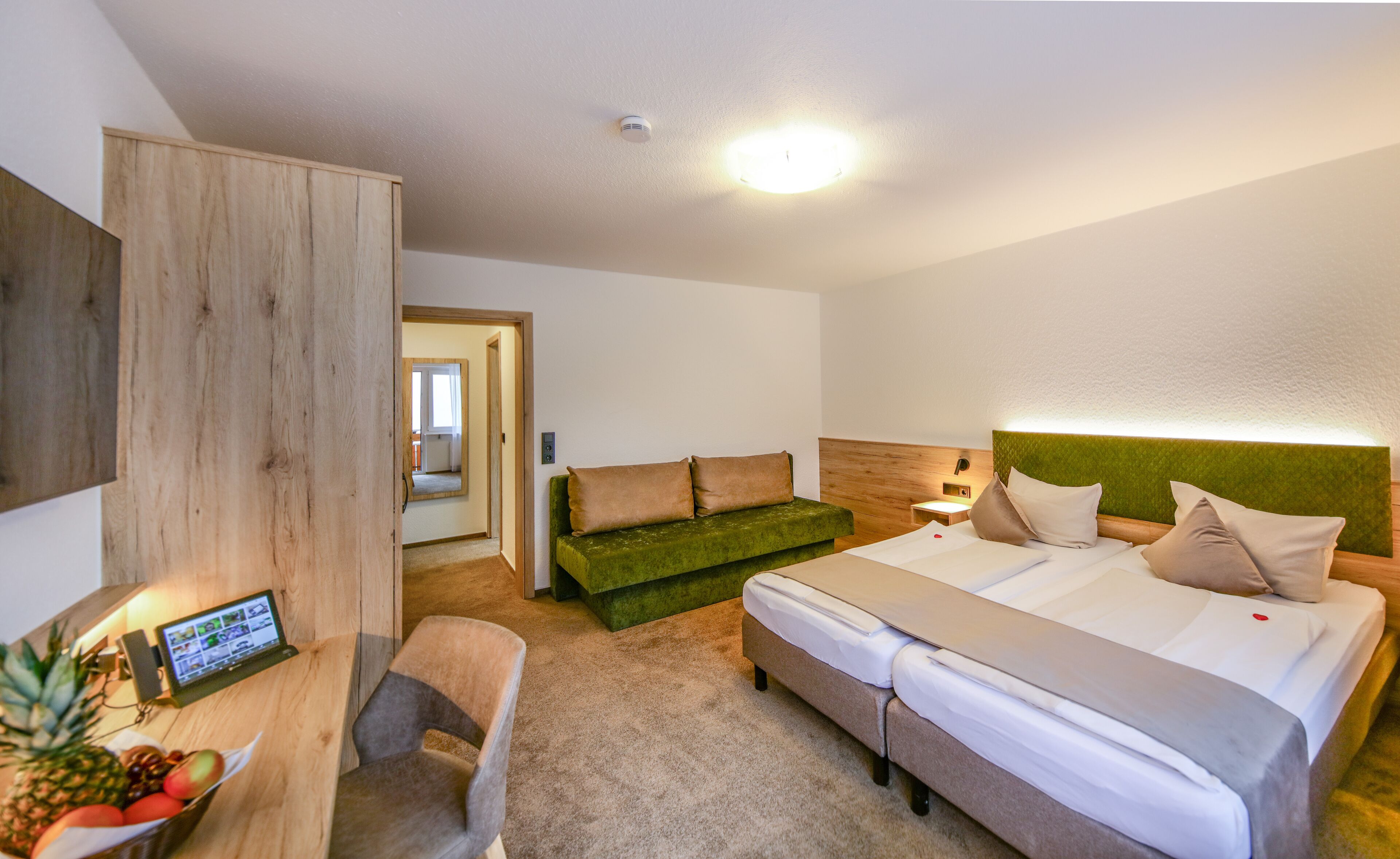 Ein Hotelzimmer mit einem Bett, einem grünen Sofa, einem Schreibtisch mit einem Computer und einem kleinen Tisch mit Früchten.