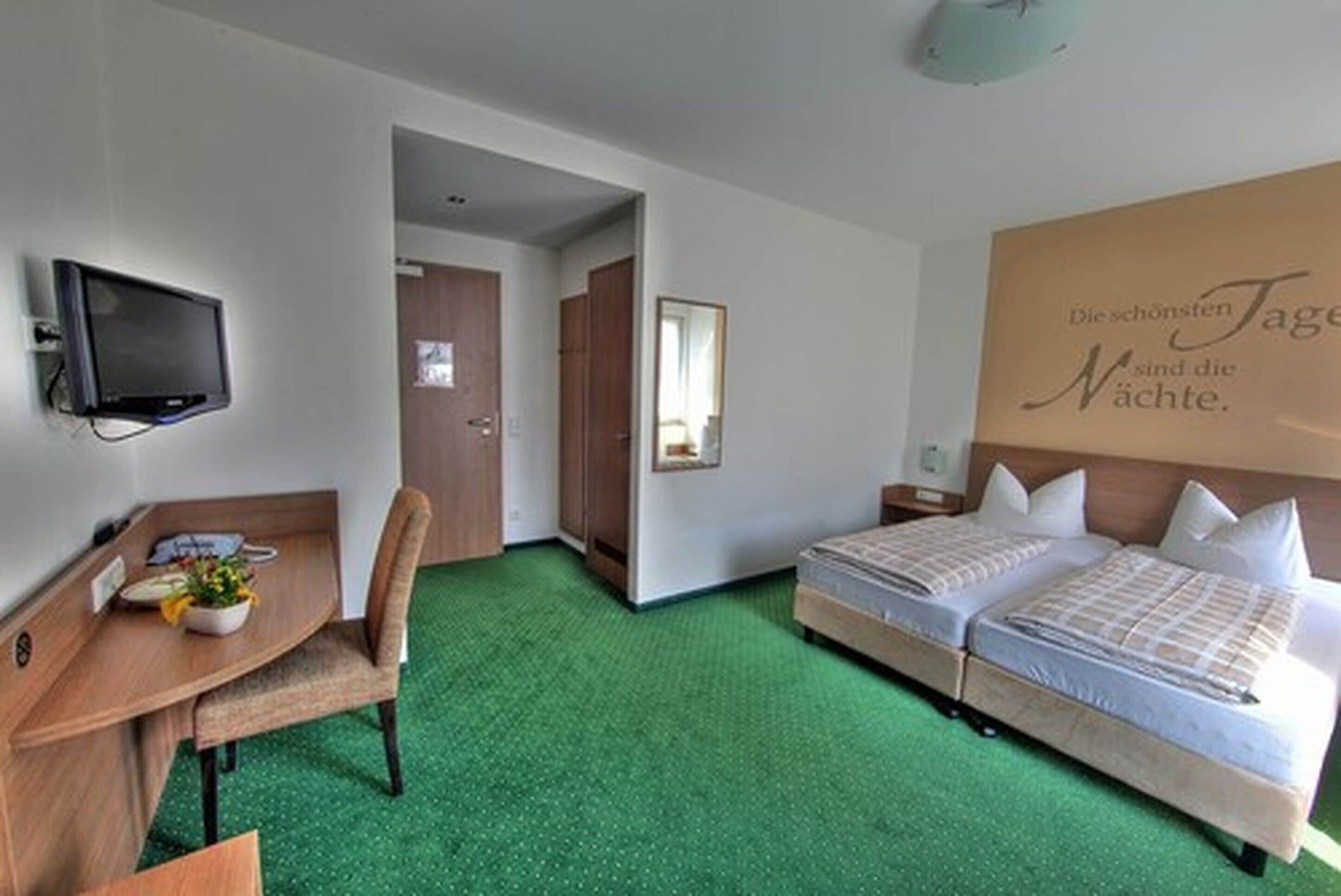 Quarto de hotel com uma cama, escrivaninha, cadeira, televisão e espelho.