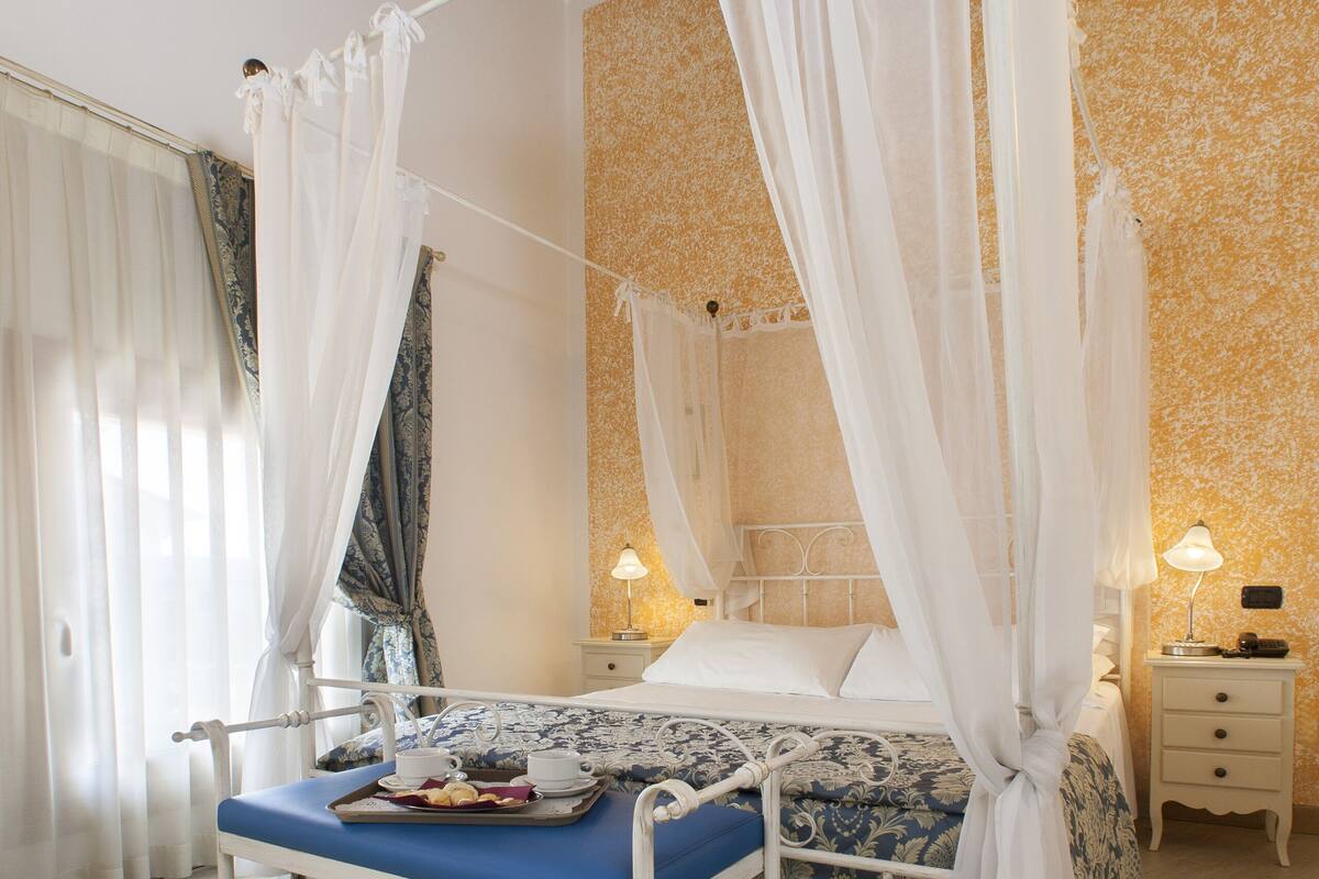A four-poster canopy bed with a tray of breakfast items, a bedside table with a lamp, and a patterned wall.