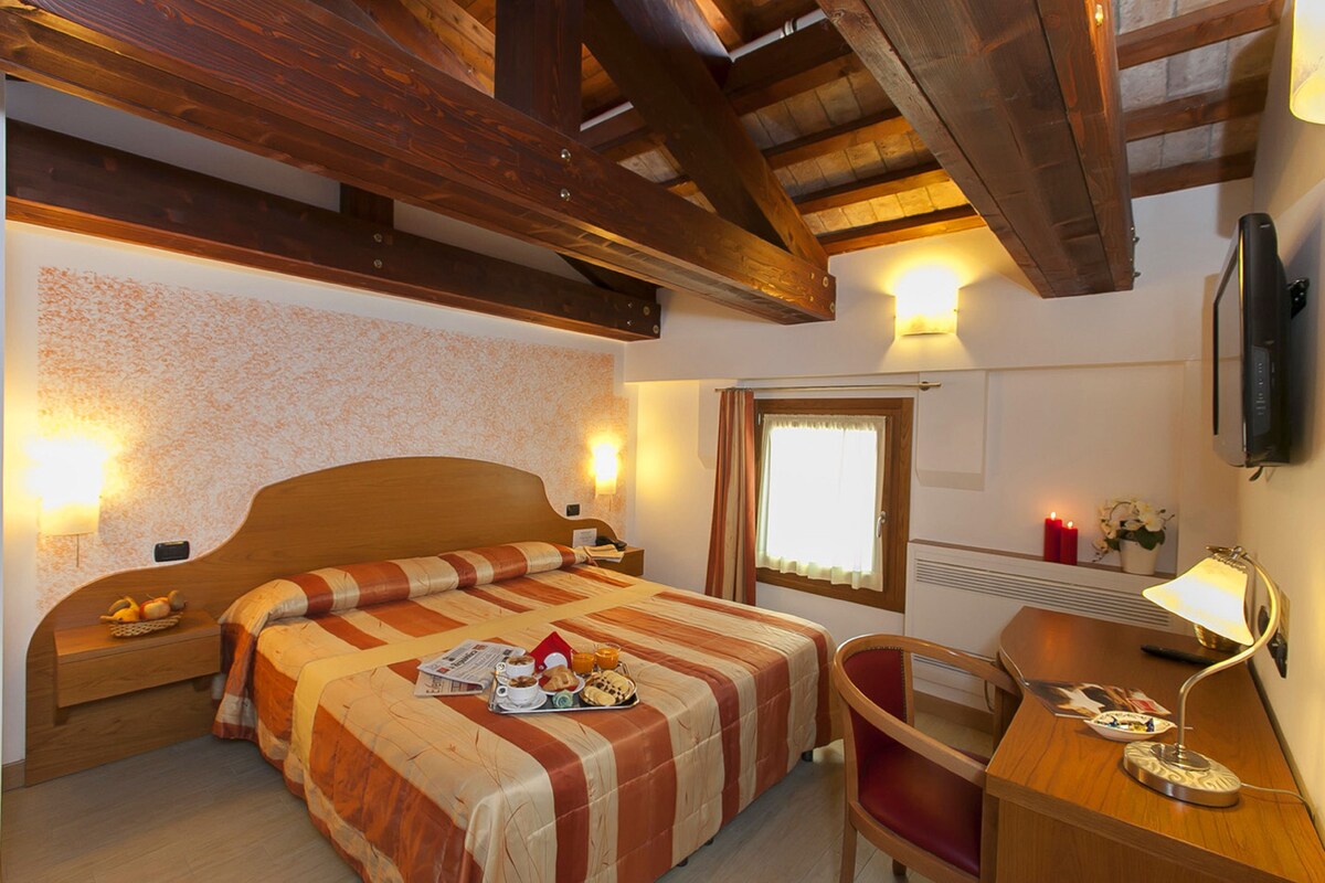 A bedroom with a wooden bed, bedside tables, a desk, a chair, a lamp, a window with curtains, and a wall-mounted TV.