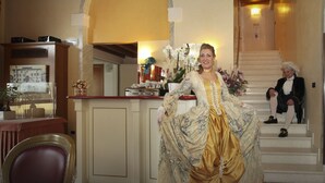 Una persona vestida con un traje histórico en una habitación que tiene una escalera, un comedor y una cocina al fondo.