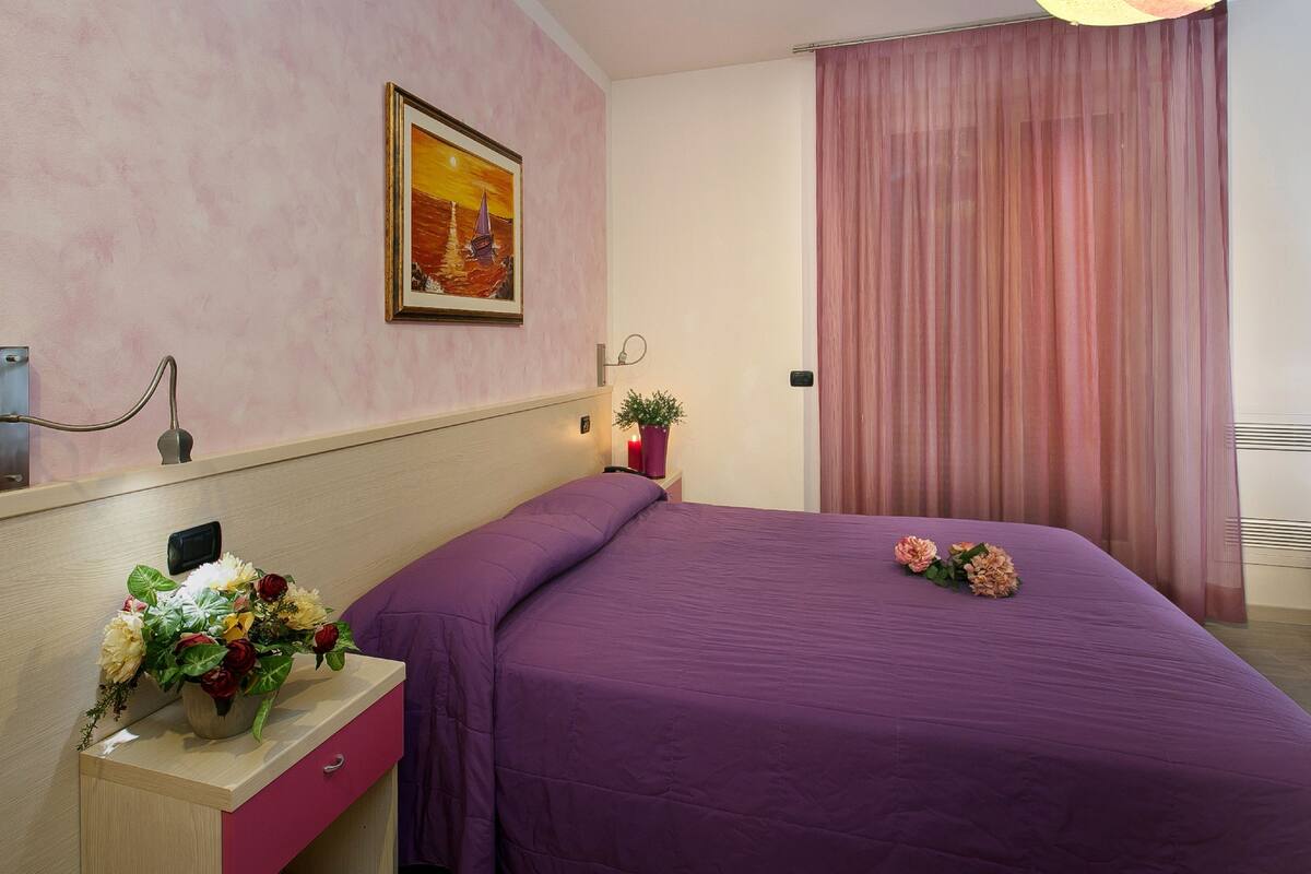 A hotel room with a purple bedspread, a nightstand with a vase of flowers, a painting on the wall, and a window with pink curtains.