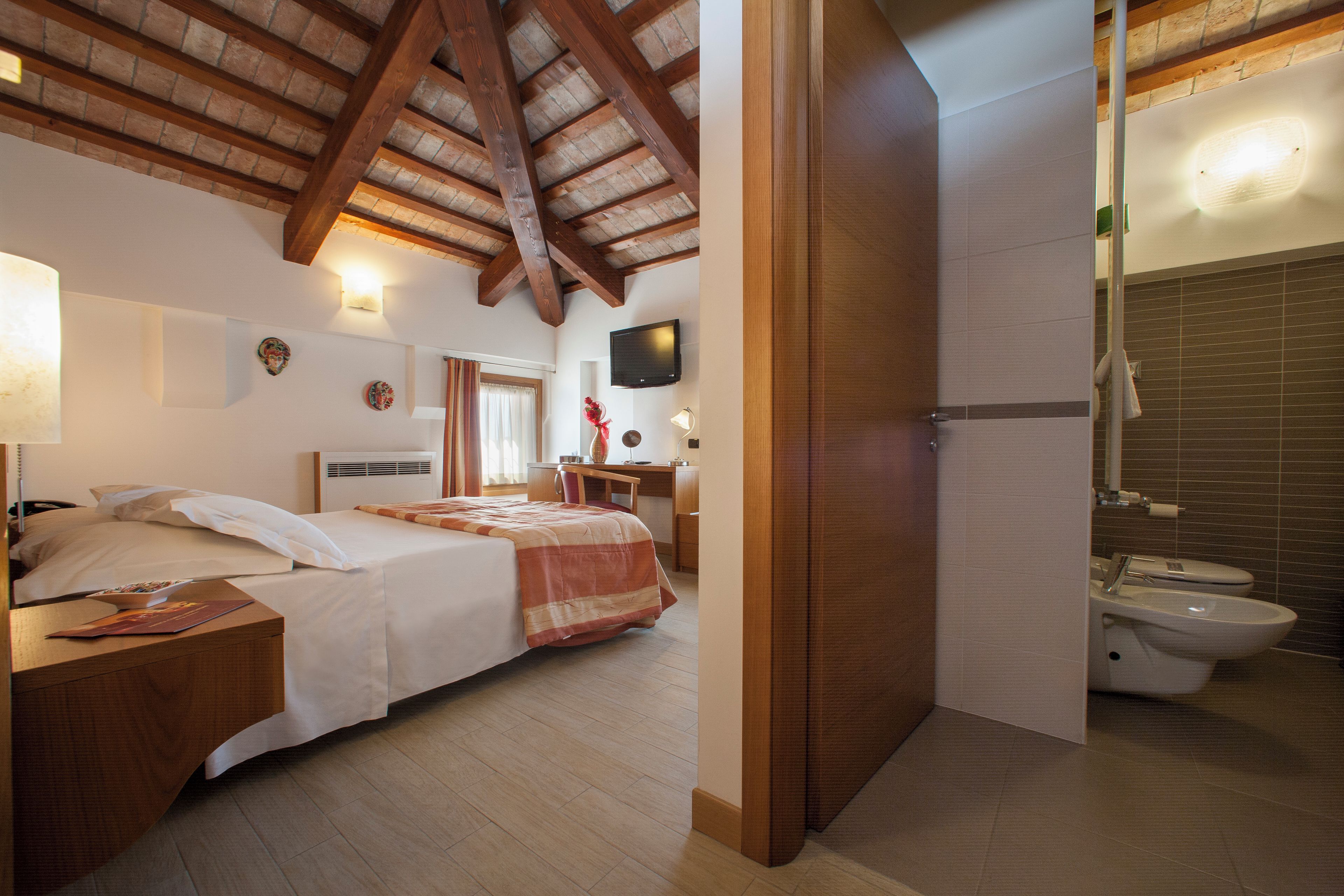 A hotel room with a wooden ceiling, a bed with white linens, a desk with a chair, a television on the wall, and a bathroom with a toilet and sink.
