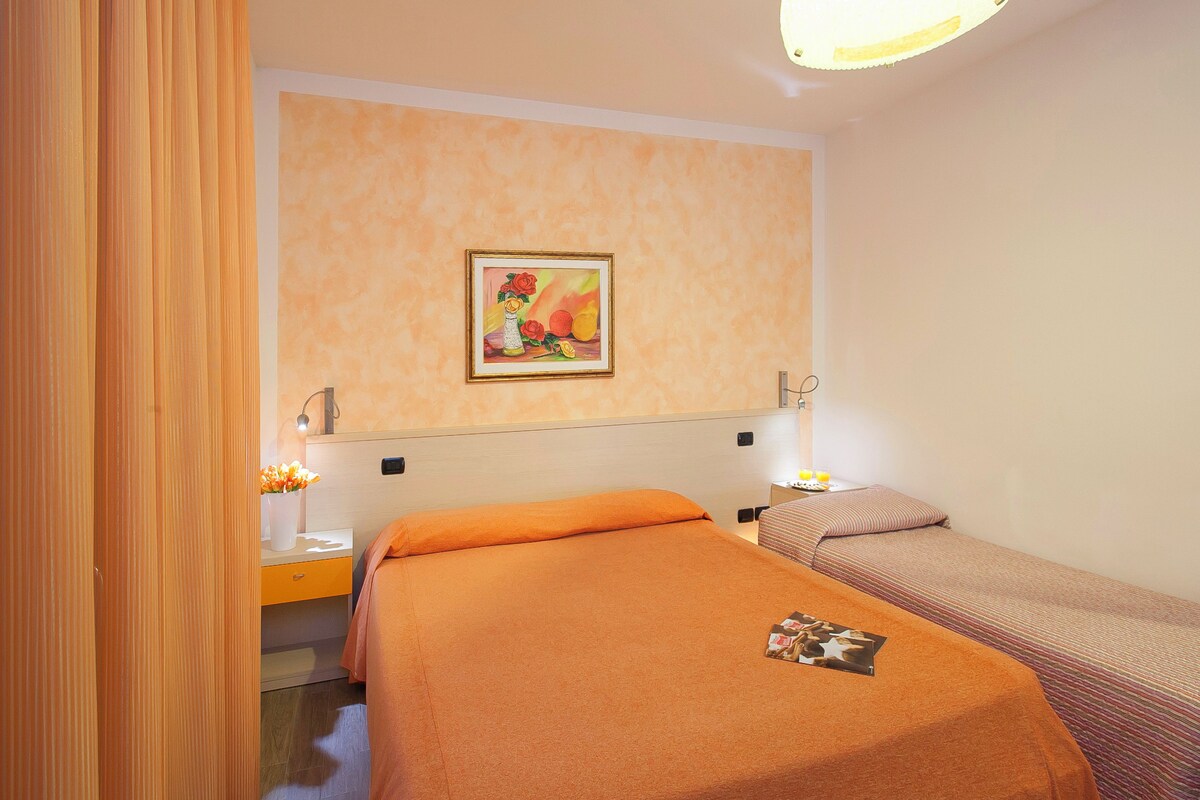 A hotel room with a bed, a bedside table, a nightstand, a lamp, and a painting on the wall.
