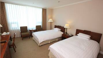 A hotel room with two beds, a desk with a chair, a small table, and a lamp.