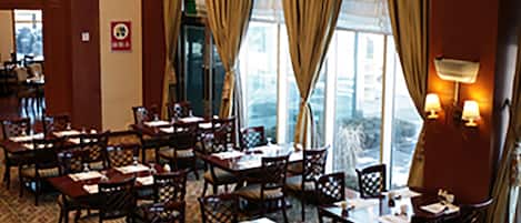 A hotel dining area with multiple tables and chairs, large windows with curtains, and wall-mounted lamps.