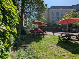 A grassy outdoor area with picnic tables and umbrellas, a brick building in the background, and parked cars.