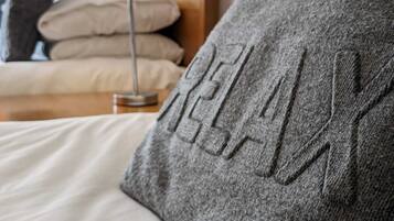 A bed with a grey pillow featuring embossed text, a bedside lamp, and neatly folded beige pillows in the background.
