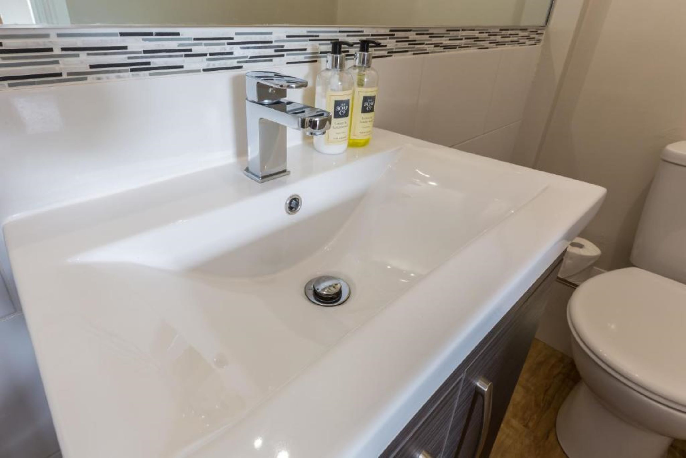 Un lavabo blanc dans la salle de bain, avec un robinet chromé, deux flacons de lotion et des toilettes.