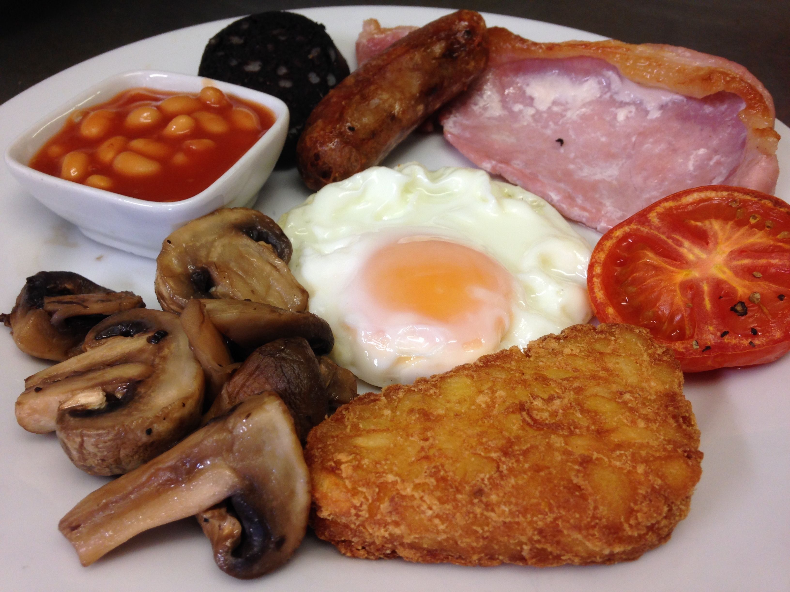A plate of breakfast food including eggs, bacon, sausages, mushrooms, baked beans, and a tomato slice.