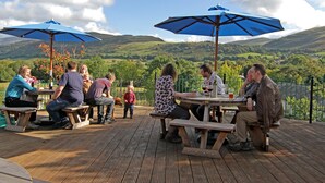 A group of people seated at wooden tables on a deck with blue umbrellas, overlooking a scenic landscape with hills and trees. - Embleton Spa Hotel (Cockermouth)