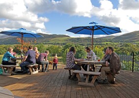 A group of people seated at wooden tables on a deck with blue umbrellas, overlooking a scenic landscape with hills and trees.