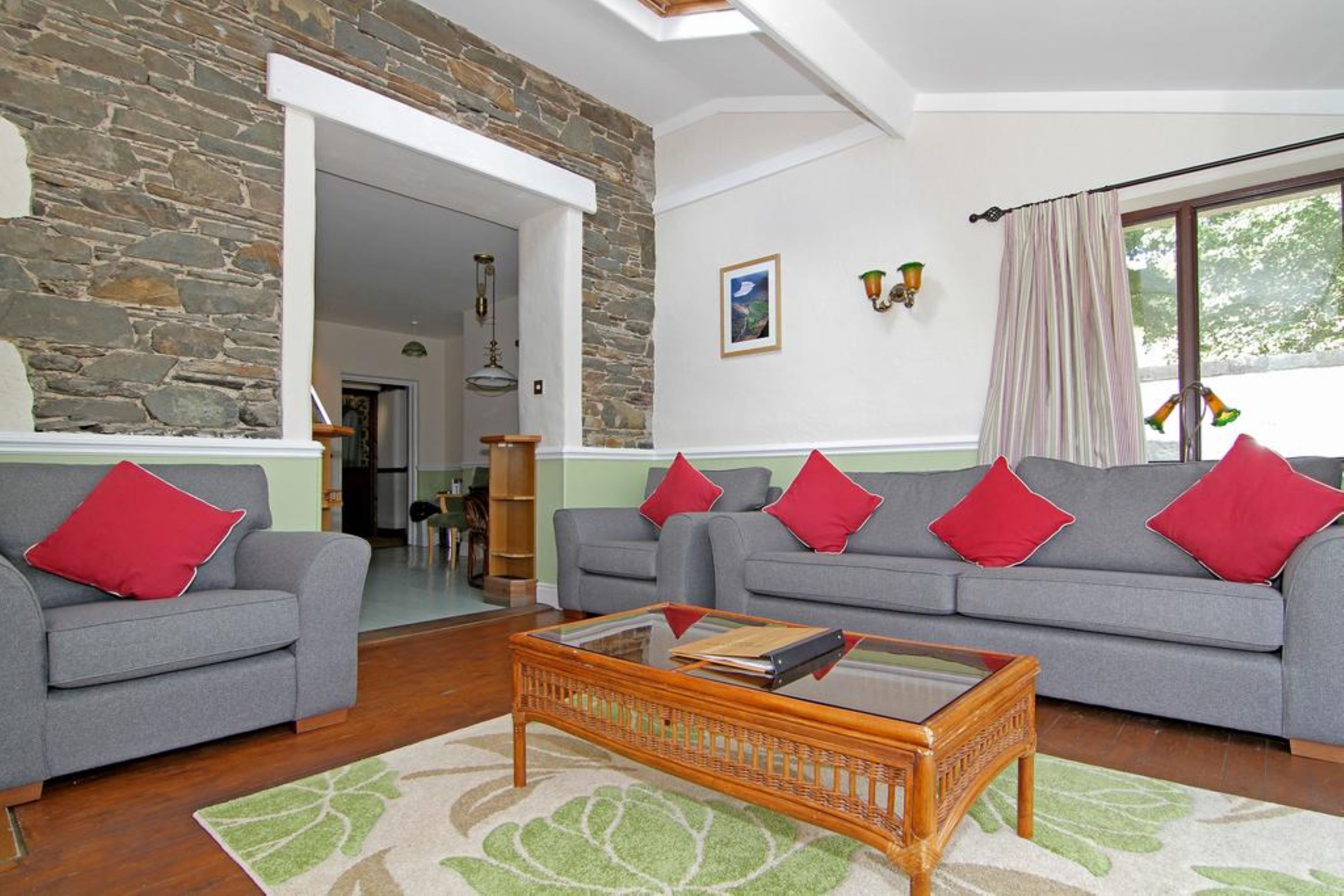 Sala de estar com parede de pedra, dois sofás cinzentos com almofadas vermelhas, uma mesa de centro de vidro e vista pela janela com cortinas.