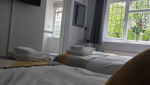 A hotel room with two beds, a window with a view of greenery, a mirror, and a small table with toiletries. - Avonlea Guest House (Banbury)