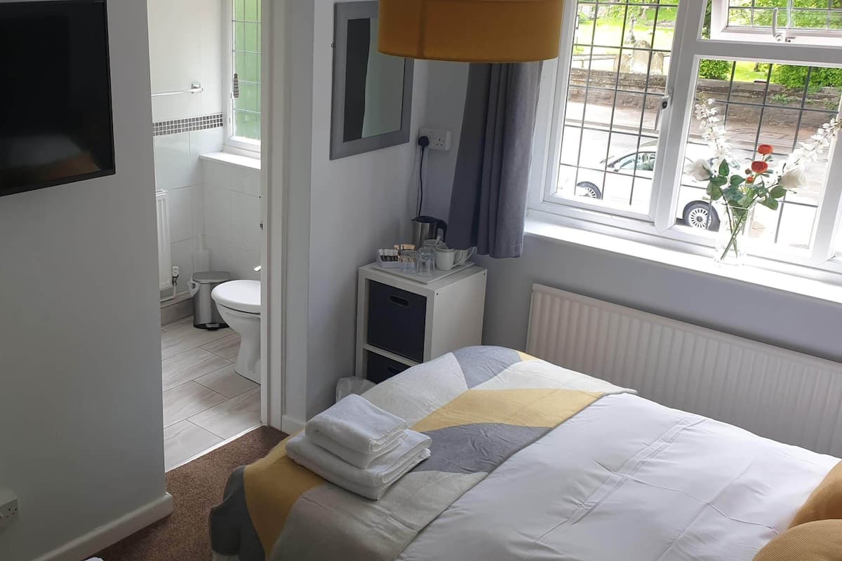 A bedroom with a bed, a TV mounted on the wall, a window with a view of the street, and a small bedside table with a lamp.