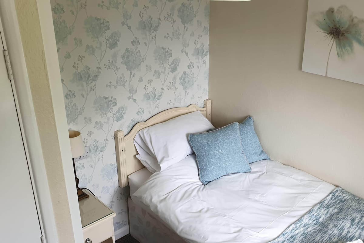 A small, neatly made bed with white bedding and a blue pillow, a nightstand with a lamp, and floral wallpaper.