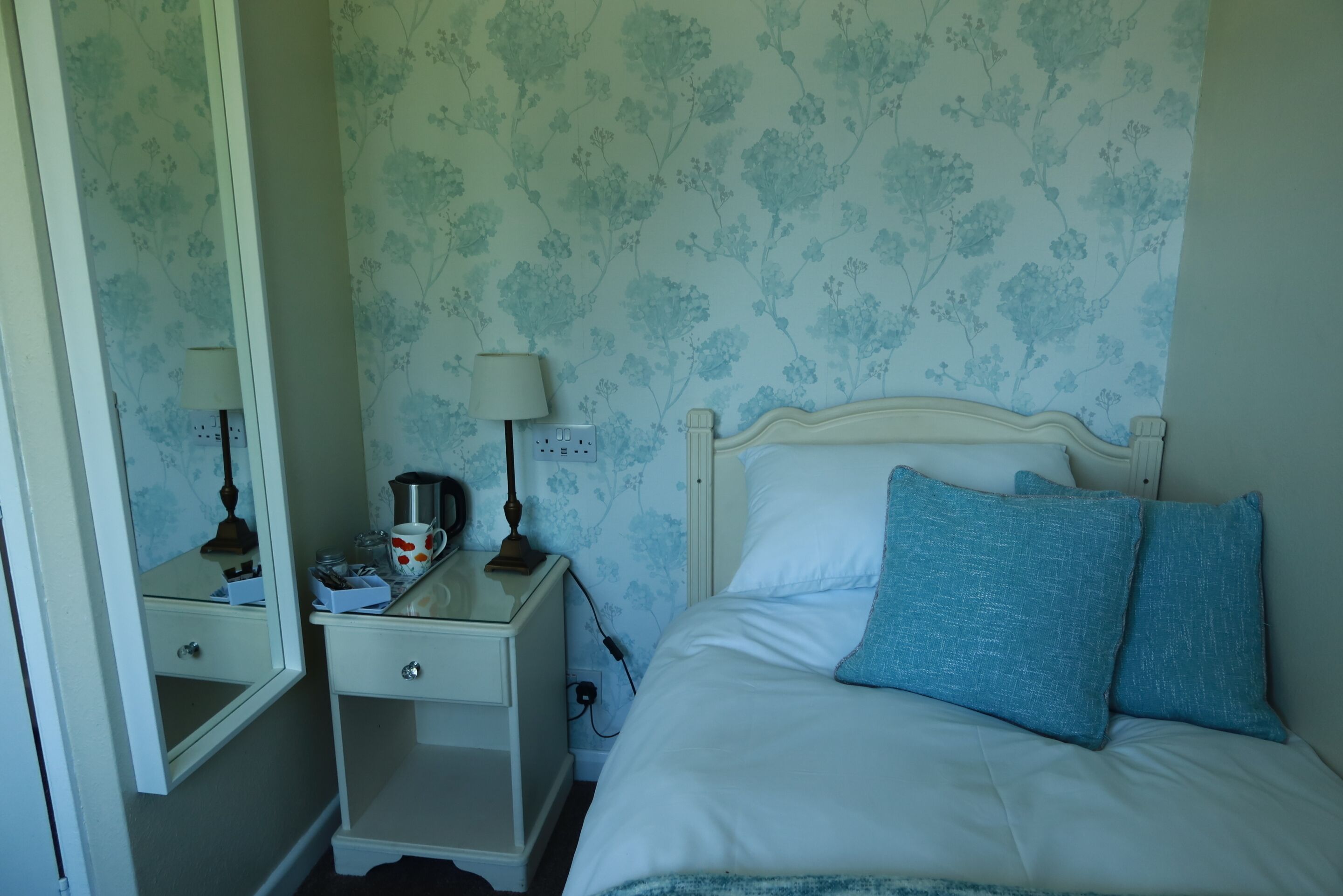 A bedroom with a bed, a nightstand, a mirror, and a wall with floral wallpaper.
