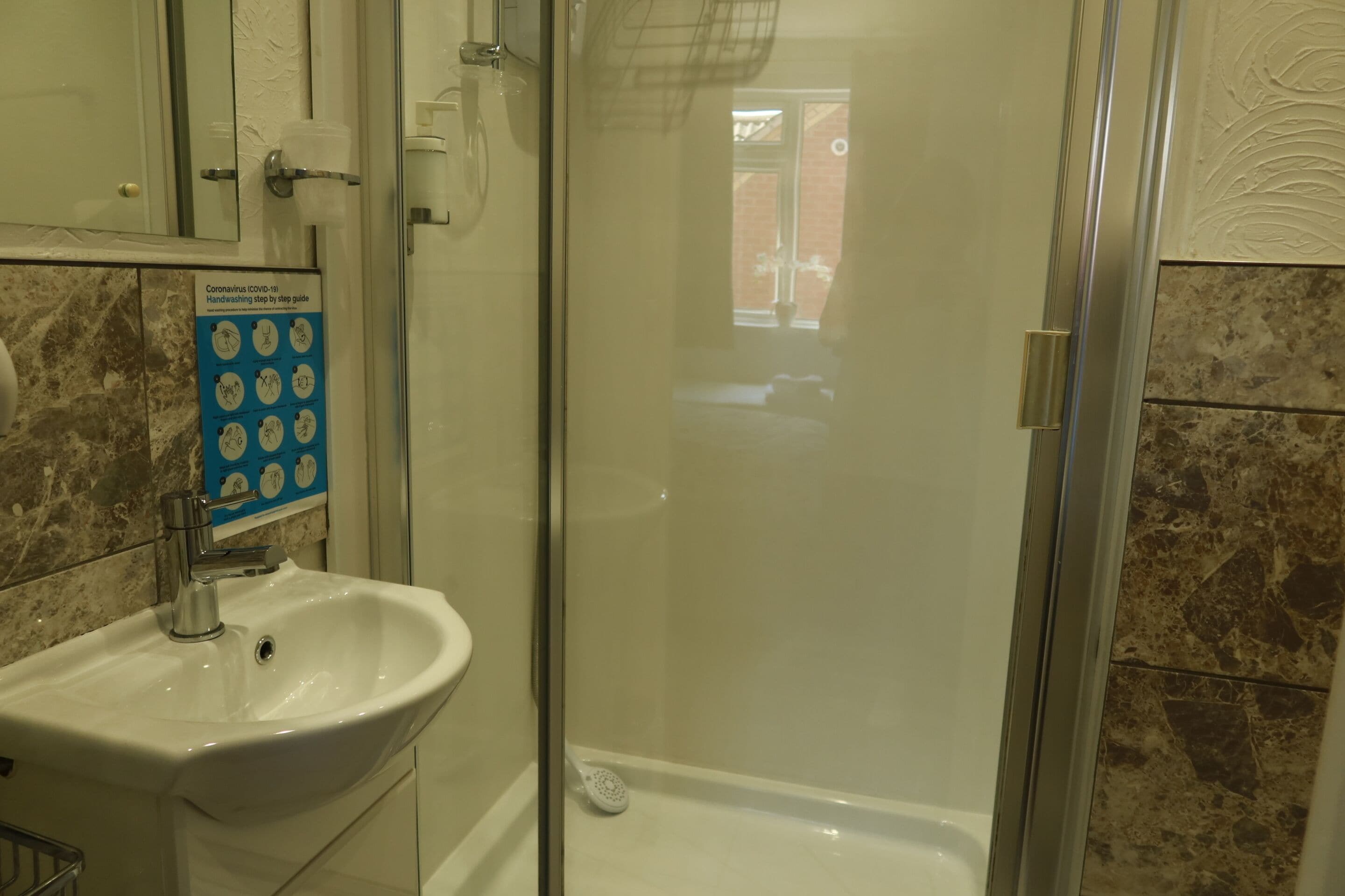 A bathroom with a white sink, a glass shower enclosure, and a handwashing guide on the wall.