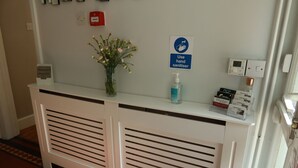 A reception desk with a hand sanitizer dispenser, a vase with flowers, and a sign instructing to use hand sanitizer. - Avonlea Guest House (Banbury)