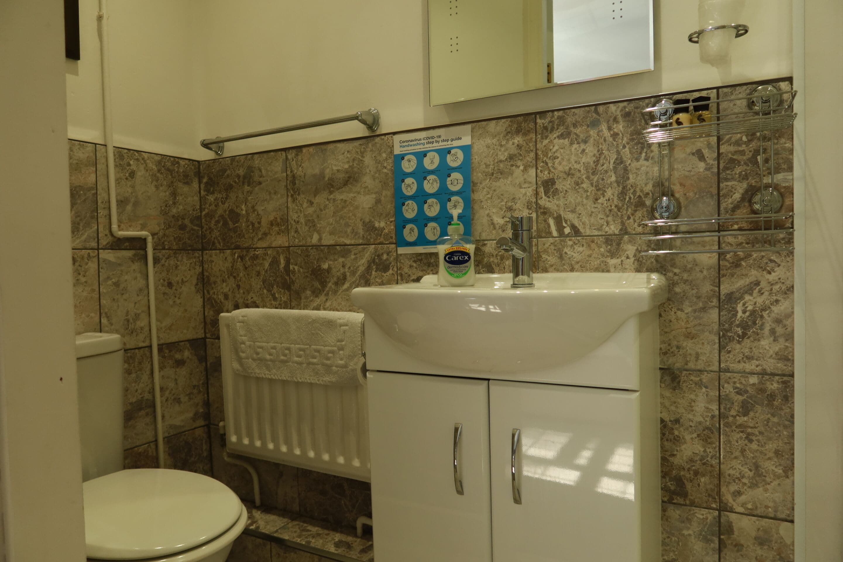 A bathroom with a white toilet, a sink with a soap dispenser, a towel rack, and a poster with COVID-19 guidelines.